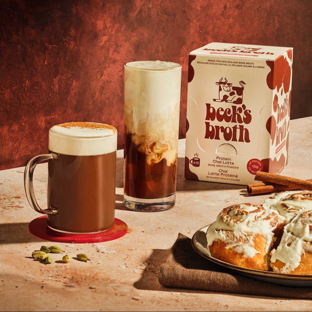 A styled product shot featuring a box of Beek's Broth Protein bone broth chai latte with foam, and cinnamon rolls on a rustic table against a textured dark red-brown background.